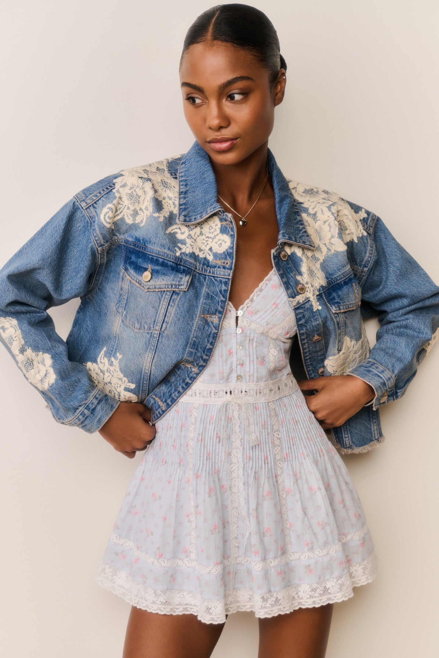 Woman wears blue denim jacket with floral lace embroidery and a light blue floral skirt on a beige background