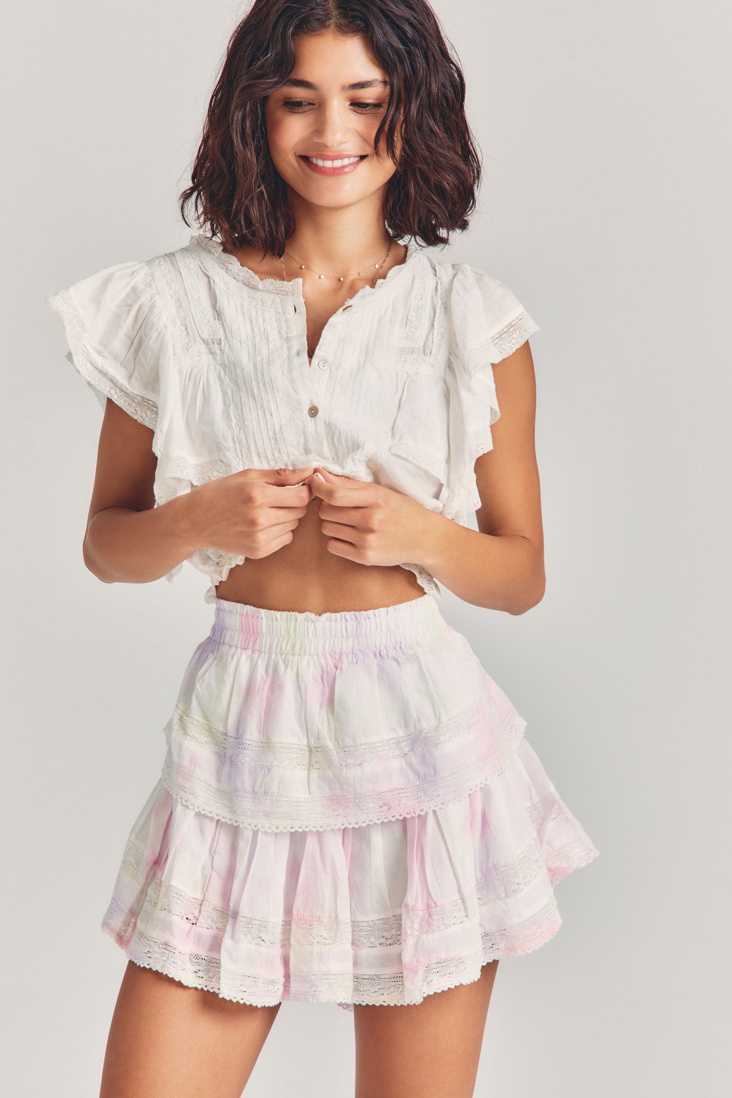Woman wears white ruffle cropped top with mini white and pink tie-dye ruffle skirt against beige background
